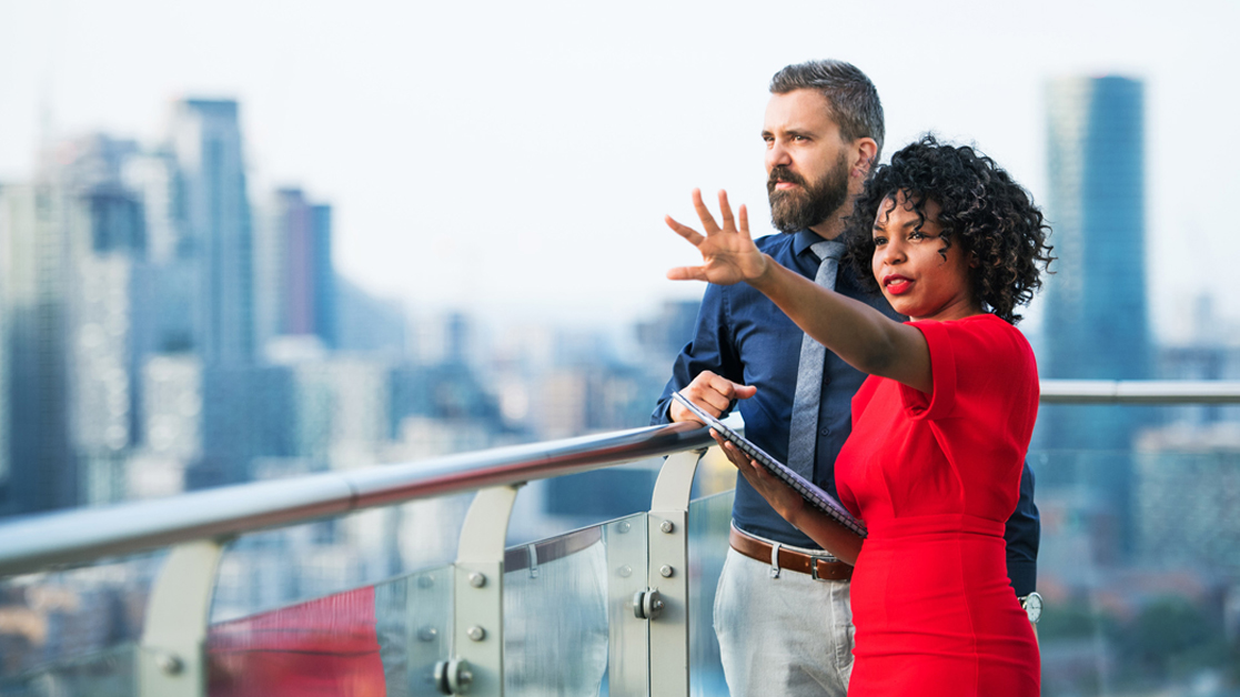 Two business professionals standing on a rooftop, looking out over a city and talking