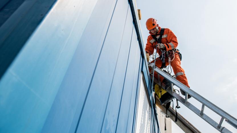 Man wearing orange high visibility gear and helmet is leaning on a rooftop from a ladder